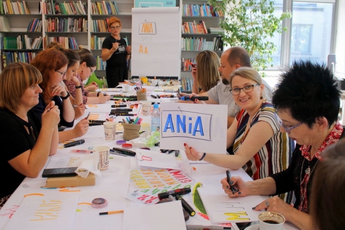 Na zdjęciu panie uczestniczące w warsztatach, siedzą przy stole - jedna z nich prezentuje kartkę ze swoim imieniem ANIA