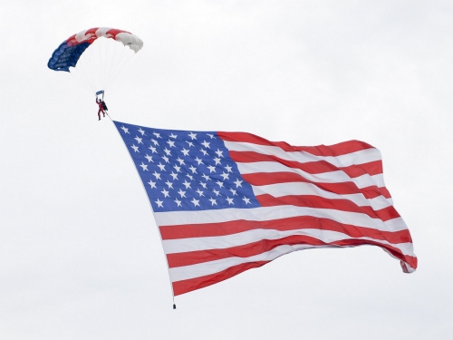 Skoczek spadochronowy nad jego głową rozłożony spadochron w barwach flagi USA ciągnie za sobą ogromną flagę USA