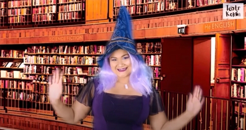Dziewczyna w kolorowych fioletowych włosach z czapką czarodzieja stoi z rozłożonymi rękami, a w tle jest dużo regałów pełnych książek