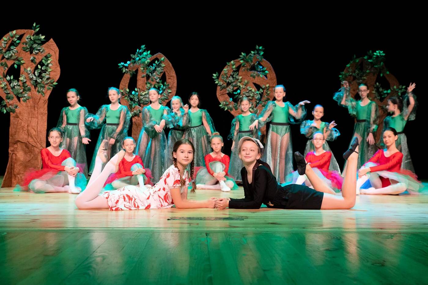 Na scenie z tyłu dziewczynki ubrane w zielone stroje baletowe przebrane za drzewa, na pierwszym planie siedzą uśmiechnięte i ubrane w czerwone stroje baletowe
