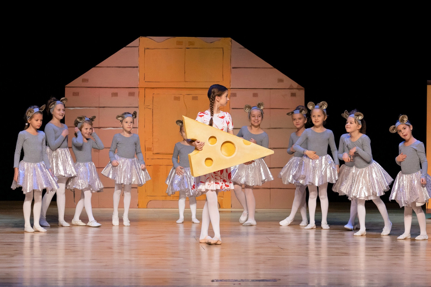 Na scenie w głównym miejscu dziewczynka trzyma ogromny kawał sera za nią duża ilość innych dziewczynek przebranych za myszy