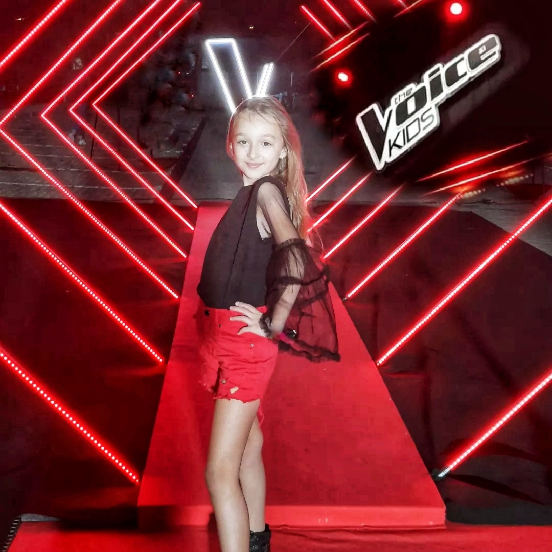 Na zdjęciu młoda Marysia Błaszczyk pozuje na tle ścianki z napisem The Voice Kids. Ubrana jest w czerwone krótkie spodenki i czarną bluzkę