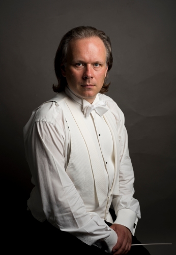 Na zdjęciu dyrygent w białej koszuli, białej kamizelce i białej muszce w ręku trzyma białą batutę