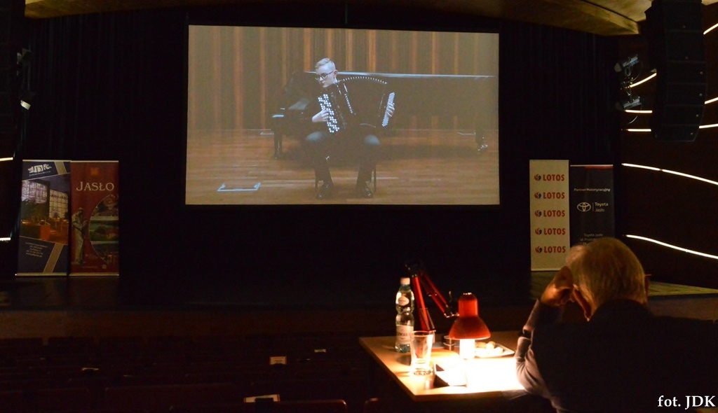 Na zdjęciu widać przesłuchanie online na dużym monitorze muzyk gra na harmonii, a w ciemnościach przy lampce nocnej siedzi juror, który słucha i ocenia