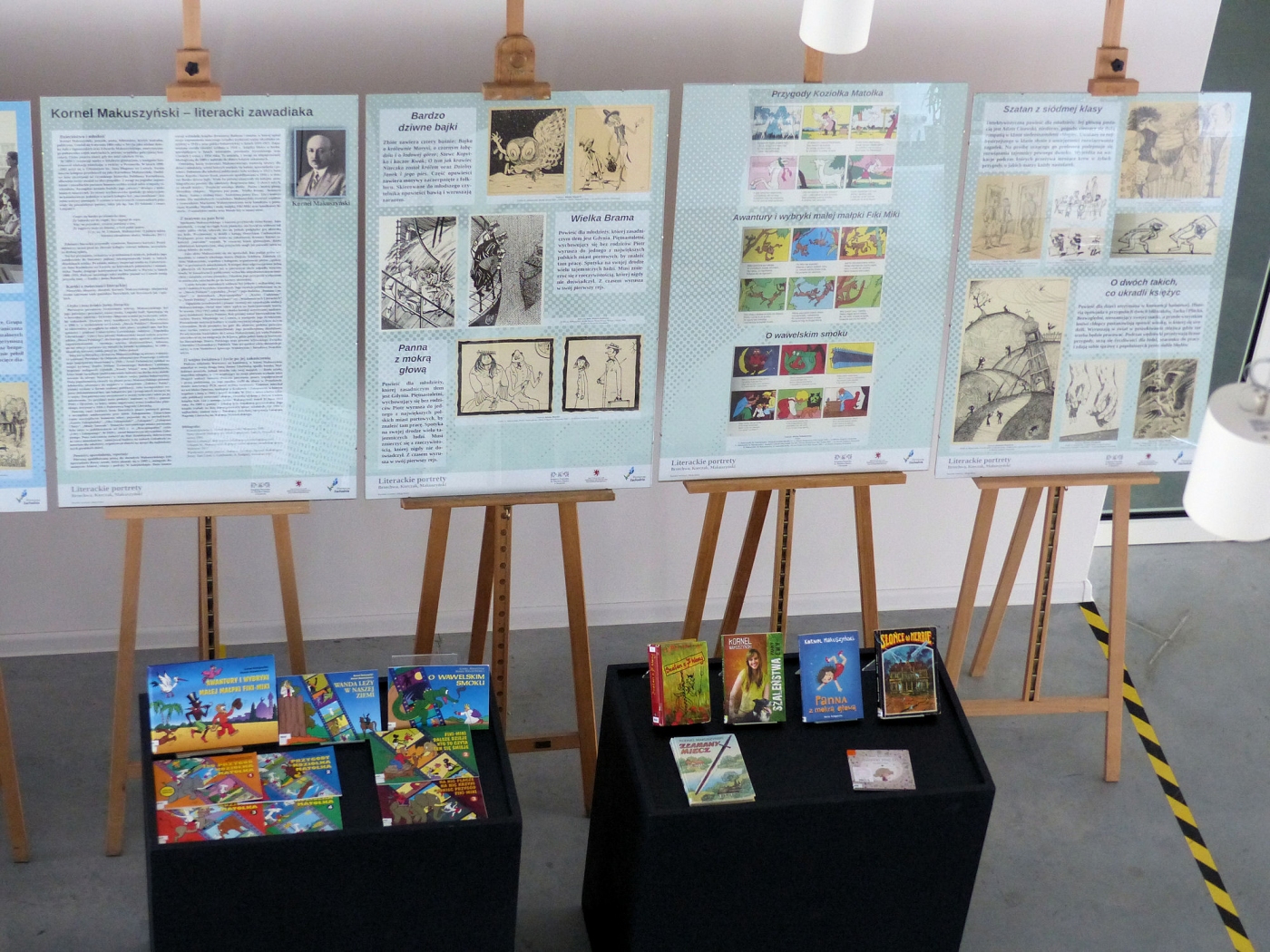 Sztalugi z informacjami dotyczącymi wystawy oraz rozłożone na stołach kolorowe okładki książek pisarzy