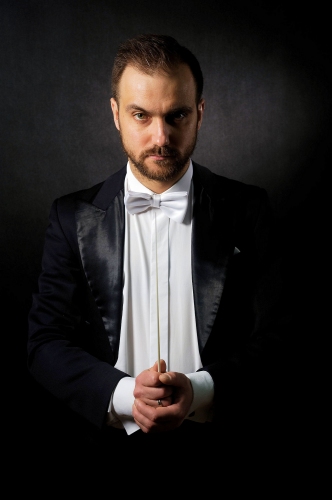 Zdjęcie portretowe, dyrygent Jakub Chrenowicz w białej koszuli, czarnym fraku i białej muszce w ręku trzyma batutę