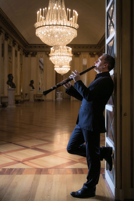 Muzyk oparty o ścianę gra na klarnecie. W tle widać rozświetlony okazały żyrandol oraz po bokach sali eksponaty popiersia nieznanych osób