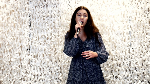 Karolina Polak śpiewa do mikrofonu. Ubrana jest w granatowo białą sukienkę.