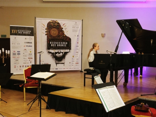 Muzyczka gra na fortepianie, w tle widać pulpit z nutami oraz ściankę z logiem festiwalu Bieszczady bez granic