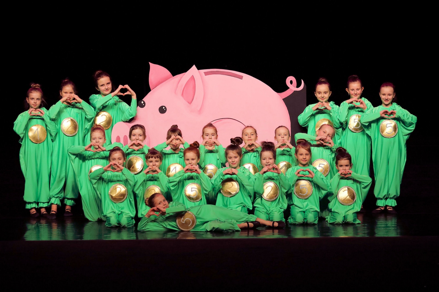 Ubrane w zielone stroje dziewczynki stoją na scenie na tle dużej różowej świnki. Ręce mają ułożone w serce