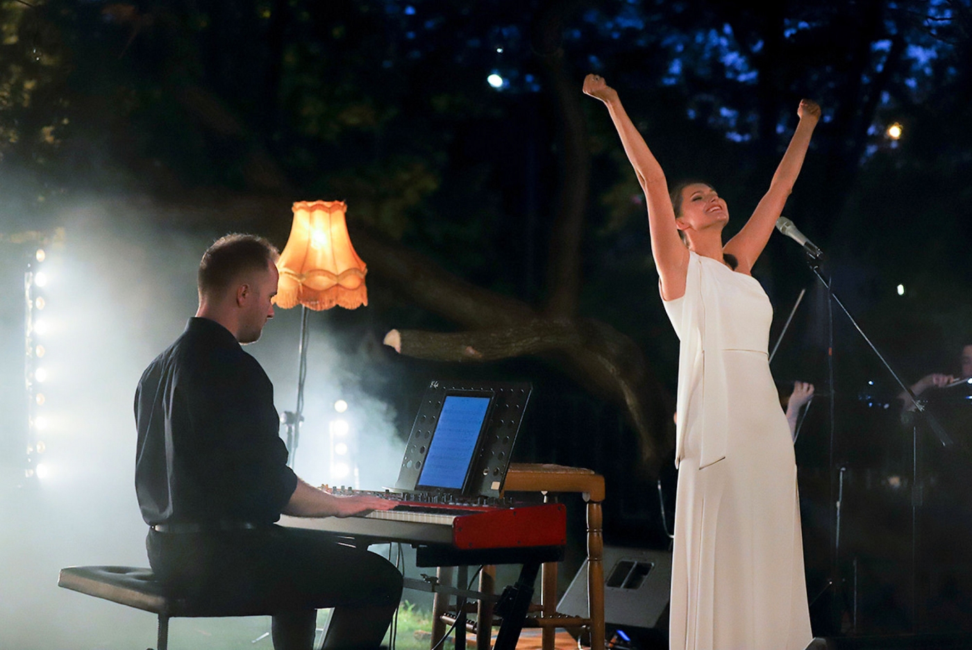 W tle widać nocne niebo na pierwszym planie piosenkarka Joanna z uniesionymi rękami w geście radości obok muzyk na kibordzie gra