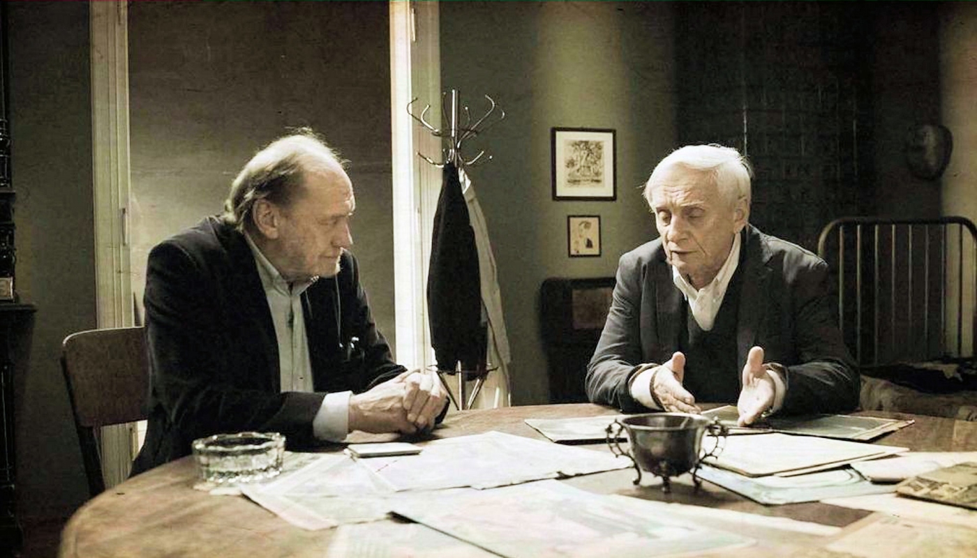 Kadr z filmu: przy stole pełnym dokumentów rozmawiają dwa aktorzy