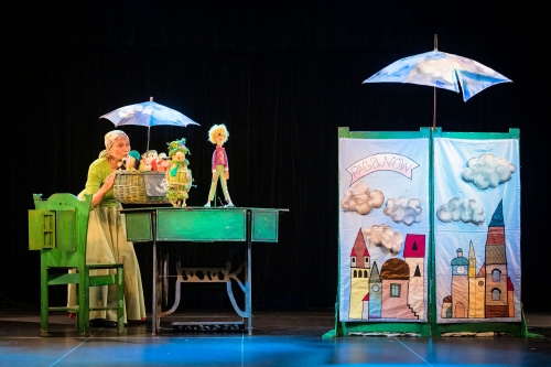 Scena ze spektaklu namalowane budynki pacanowa oraz pani i lalki oraz kolorowe parasole 