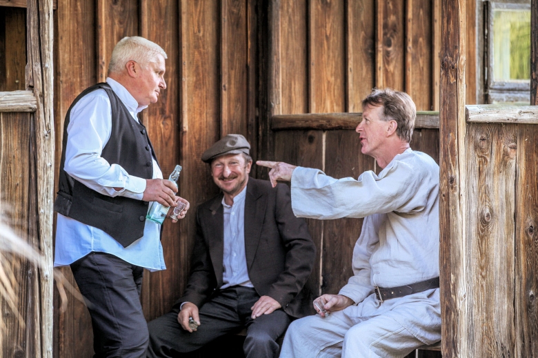 Trzech Panów w stroju ludowym przed drewnianą chatą rozmawiają, jeden trzyma butelkę drugi kieliszek