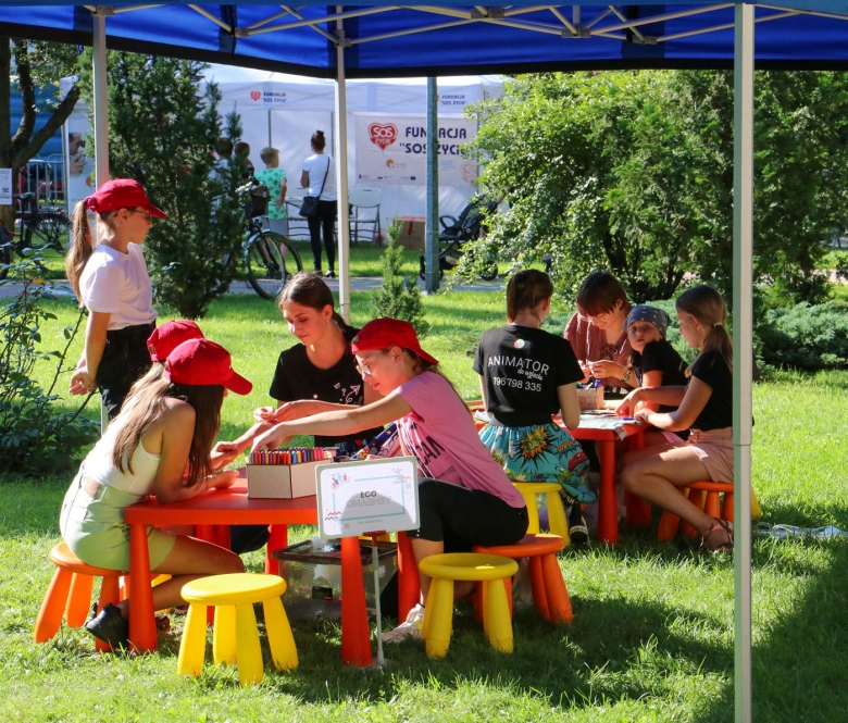 Dzieci siedzą na zielonej trawie na kolorowych krzesełkach przy stoliku wraz z animatorami