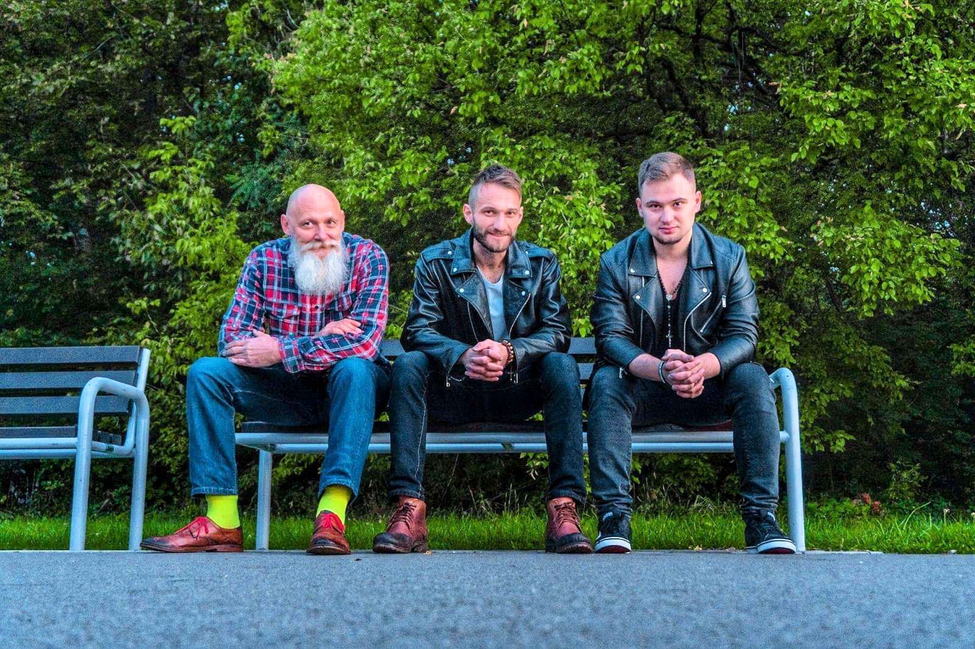 Trzech panów muzyków siedzą na ławce w parku za nimi widać zieloną trawę i zielone drzewa