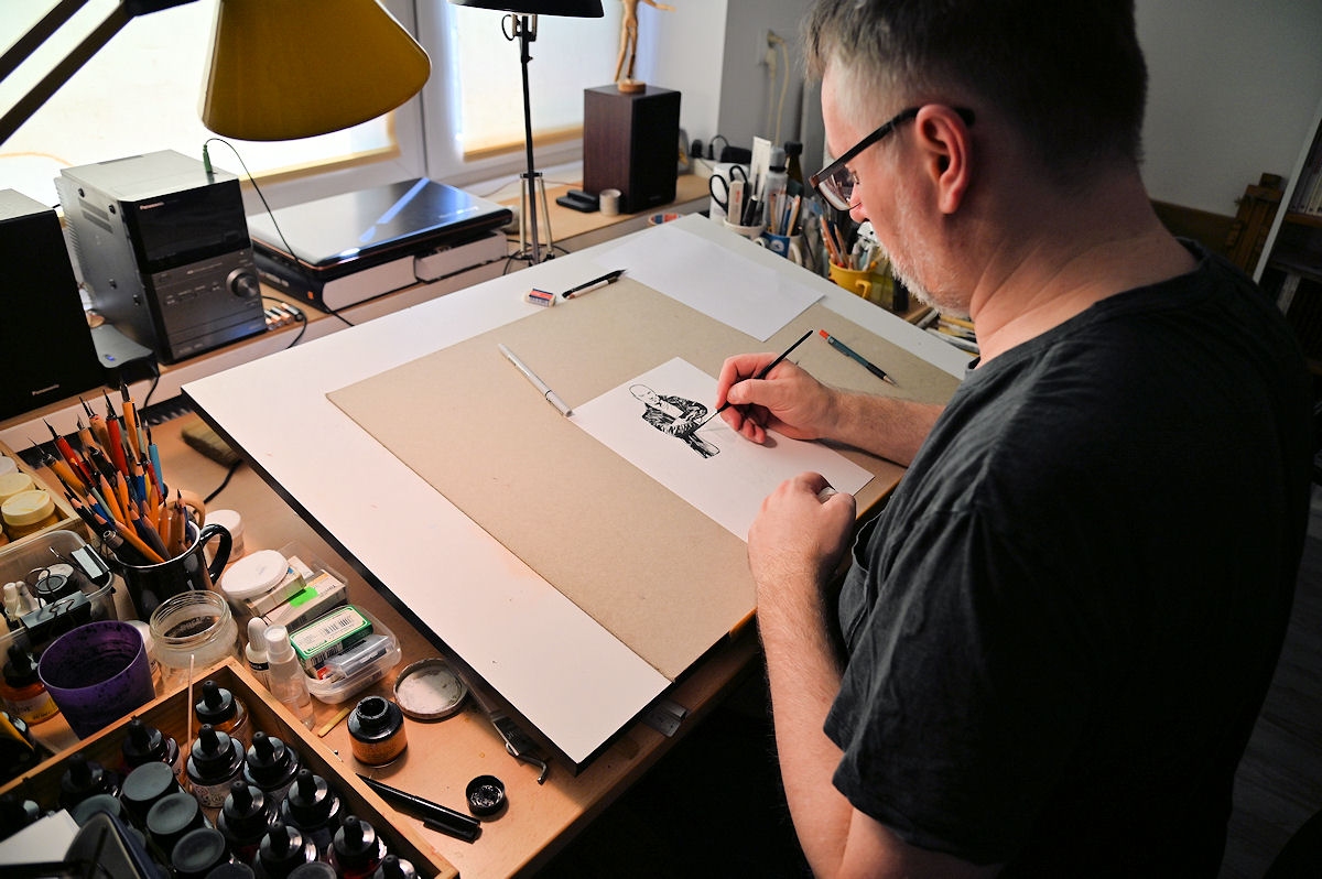 Maciej Mazur przy desce kreślarskiej w swojej pracowni rysuje czarną postać komiksową tuszem