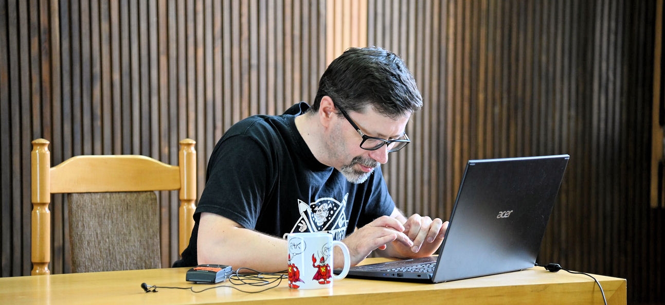 Jarek Ejsymont patrzy w ekran laptopa, obok stoi kubek z czerwonym uczłowieczonym rakiem logiem Rzeszowskiej Akademii Komiksu