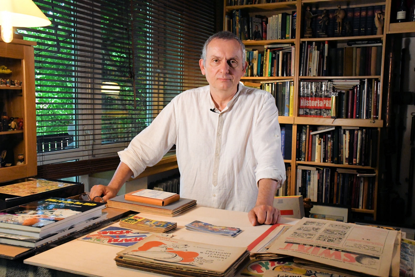 Wojciech Jama stoi w swoim mieszkaniu przed biurkiem pełnym różnych komiksów