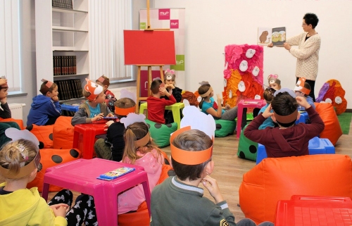 Kolorowa sala biblioteki. Dzieci siedzą i obserwują pani pokazuje im książkę z Puchatkiem