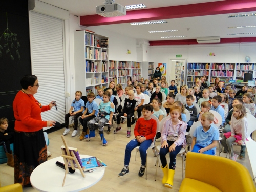 Autorka książek przemawia do sali pełnej dzieci siedzących na krzesłach w bibliotece