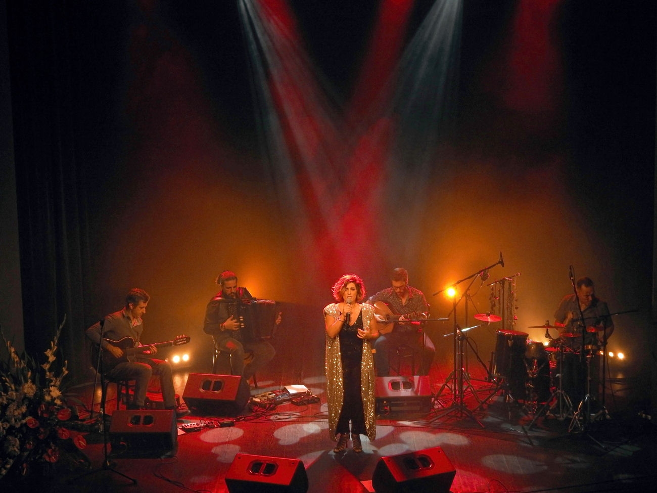 Śpiewaczka na scenie czerwone światło oświetla scenę obok reszta zespołu muzycznego siedzi na scenie i gra