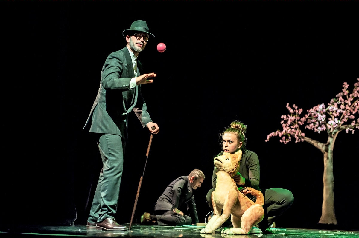 Zdjęcie, scena ze spektaklu. Pan w meloniku i z laską podrzuca piłą w tle widać psa i kucającą do niego dziewczynę