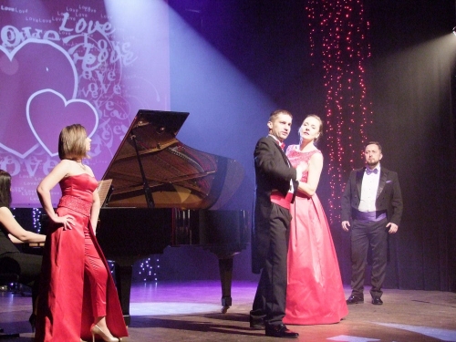 Ta sama scena ale tym razem na scenie są dwie pary dwie panie w czerwonych sukienkach i dwóch panów śpiewaków w czarnym fraku. 