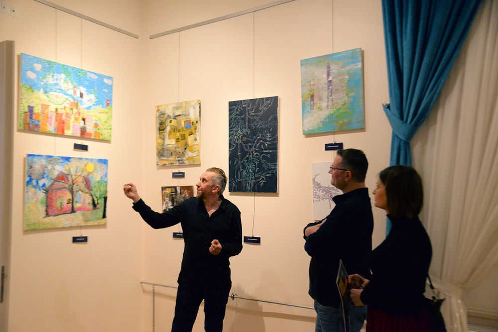 Trzy osoby na wystawie prac autora. Jedna z nich pokazuje ręką jeden z wiszących na ścianie obrazów.