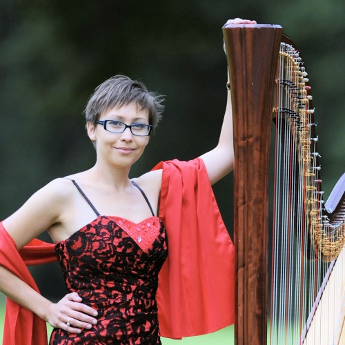 Harfistka Paulina Maciaszczyk pozuje w czerwono czarnej sukience stojąc obok dużej harfy.