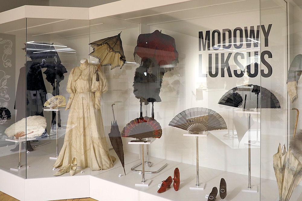 Na zdjęciu witryna wystawy. Za szklaną szybą widać zbiory: kremową suknię z epoki, torebki, kolorowe wachlarze, parasolki i damskie buty