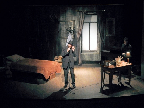 Ten sam ciemny pokój, aktor w kapeluszu mówi do mikrofonu stojącego na statywie. Po prawej stronie biurko w tle widać łóżko