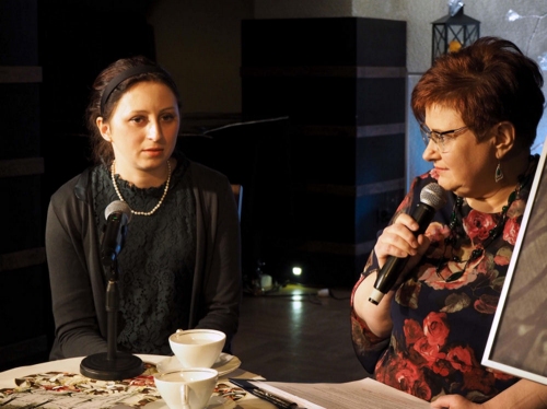 Anna Gołojuch z prowadzącą spotkanie Jolantą Nowak siedzą przy stoliku. Prowadząca spotkanie trzyma mikrofon.