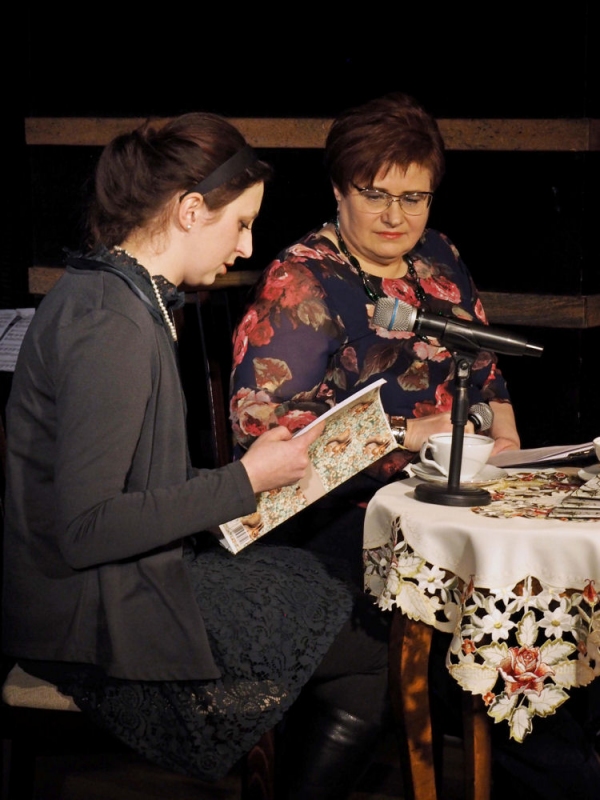 Od lewej Anna Gołojuch z Jolantą Nowak siedzą w czasie spotkania autorskiego przy stoliku. Pani Anna prezentuje zawartość książki, którą trzyma w rękach.