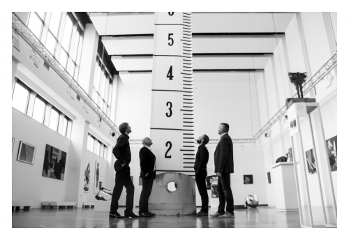 Czarno-białe zdjęcie. Czterech Panów stoi na przeciwko siebie patrzą w górę bo pośrodku sali stoi wielka linijka