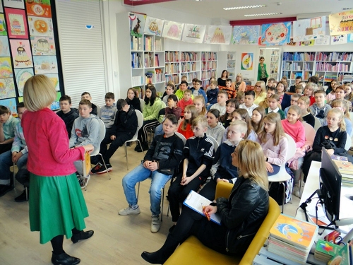 W różowej marynarce i zielonej spódnicy pisarka stoi przed salą pełną dzieci w trakcie wystąpienia