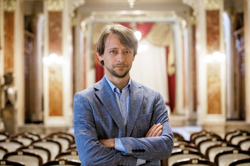 Ivan Cherednichenko – dyrygent stoi w niebieskiej marynarce na tle sali koncertowej