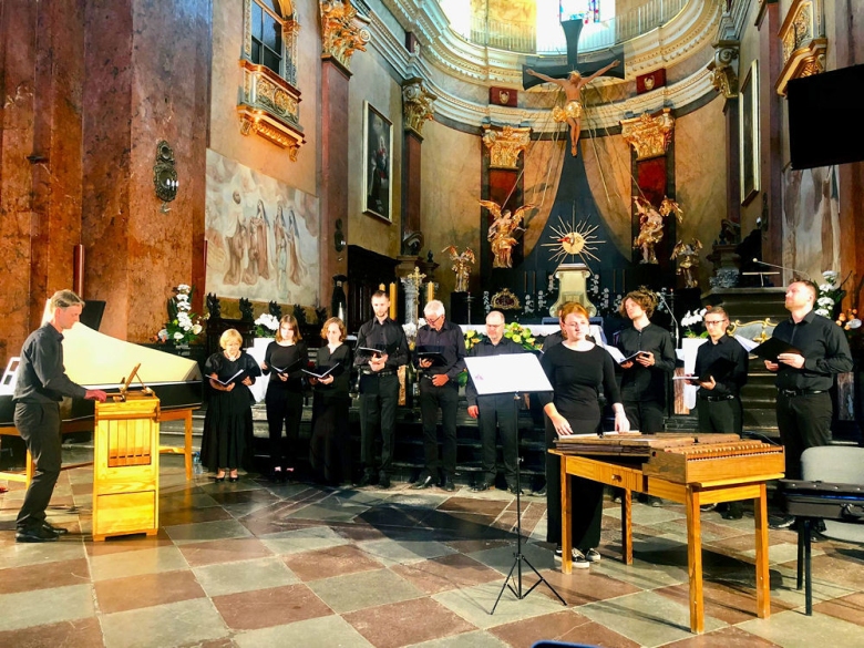 We wnętrzach kościoła koncertuje chór męski panowie ubrani w czarne koszule i czarne spodnie w ich tle widać ołtarz kościoła i duży krzyż
