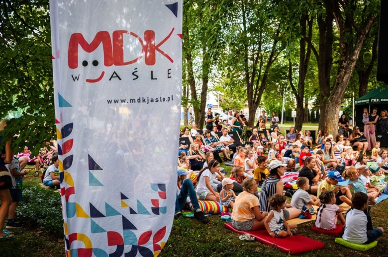Dzieci siedzą na trawie w czasie plenerowego spektaklu po lewej stronie widać baner reklamowy MDK Jasło