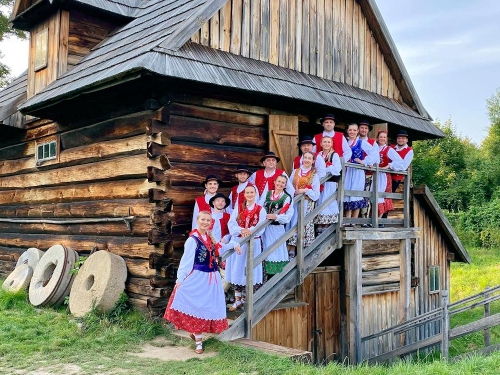 Członkowie zespołu pieśni i tańca ubrani w stroje ludowe pozują do zdjęcia na tle starego drewnianego domu